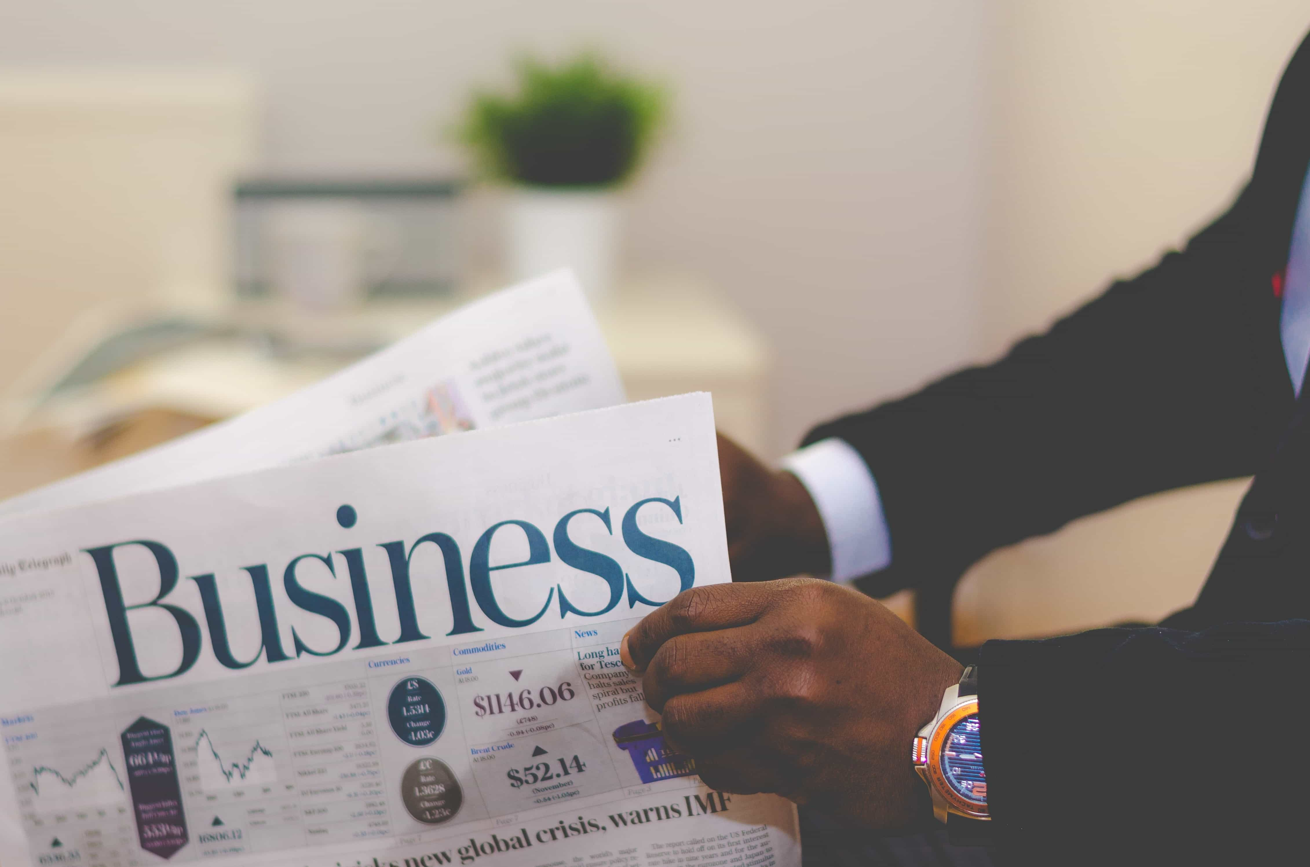 A man in a smart suit reads a business newspaper, with stocks and shares printed on the front cover.