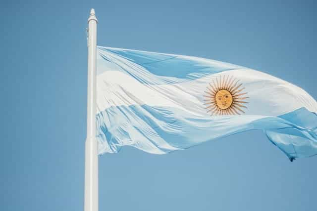 The blue and white Argentine flag waves against a blue sky, on a flagpole.