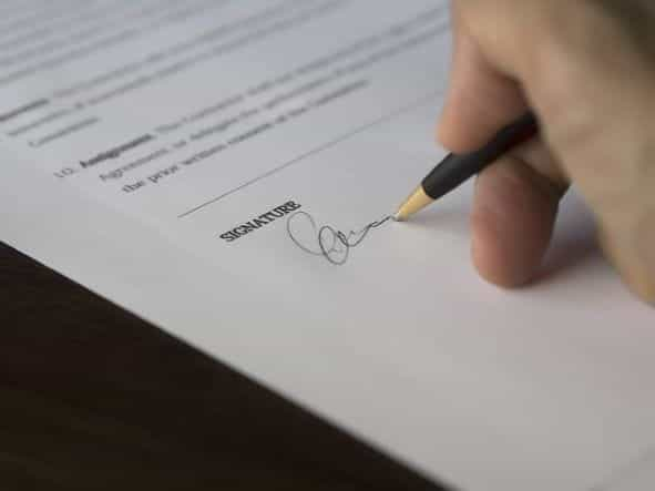 A person’s hand holding a pen and signing a written contract on paper with an official signature.