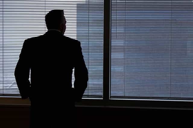 The dark silhouette of a man in a suit looking through the thin blinds of the windows in his office.