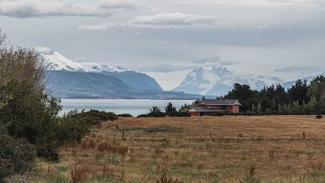 A house in a field on the coast of Puerto Natales, Chile, with snowy mountains in the distance.