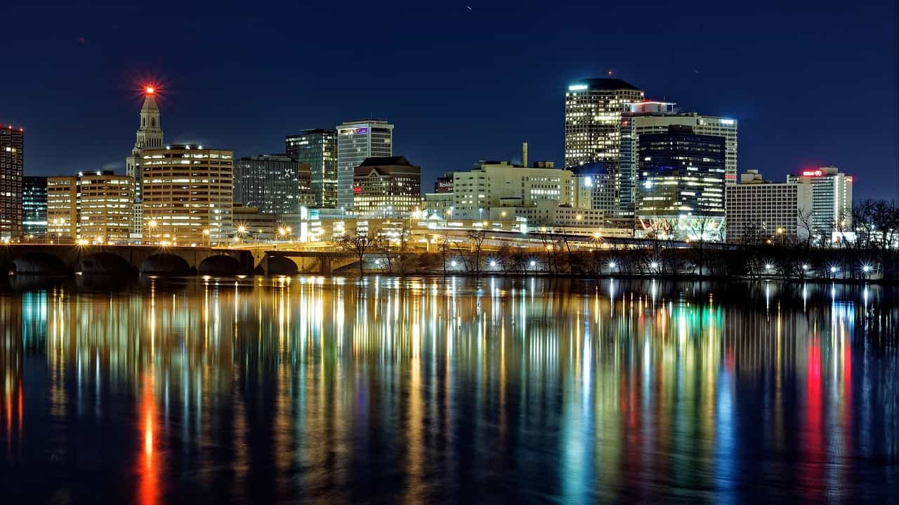 The skyline of downtown Hartford, Connecticut at night, featuring several tall, lit-up buildings whose lights are reflecting on a body of water in the foreground.