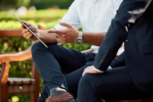 Two people in business wear sitting on a bench outside and discussing matters together, with one person showing information to the other on a tablet device.