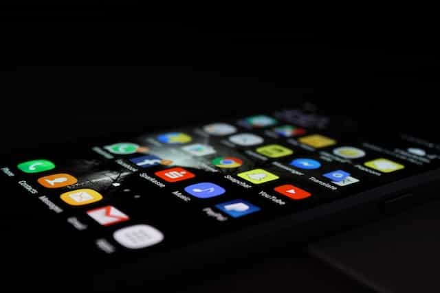 A smartphone lying flat on its back in darkness while displaying its home screen with many different mobile app icons on it.